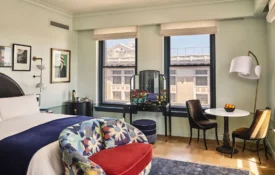 Art-filled guest room at Hotel Per La featuring plush white bedding and large windows overlooking downtown LA.