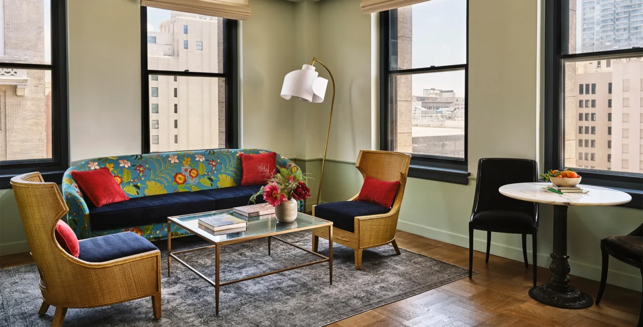 A stylish guest lounge at Hotel Per La featuring a floral sofa, wicker chairs, and windows overlooking downtown LA.