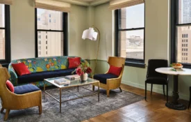 A stylish guest lounge at Hotel Per La featuring a floral sofa, wicker chairs, and windows overlooking downtown LA.