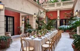 A long white banquet table is set for an outdoor dinner party in a lush, Mediterranean-style hotel courtyard.