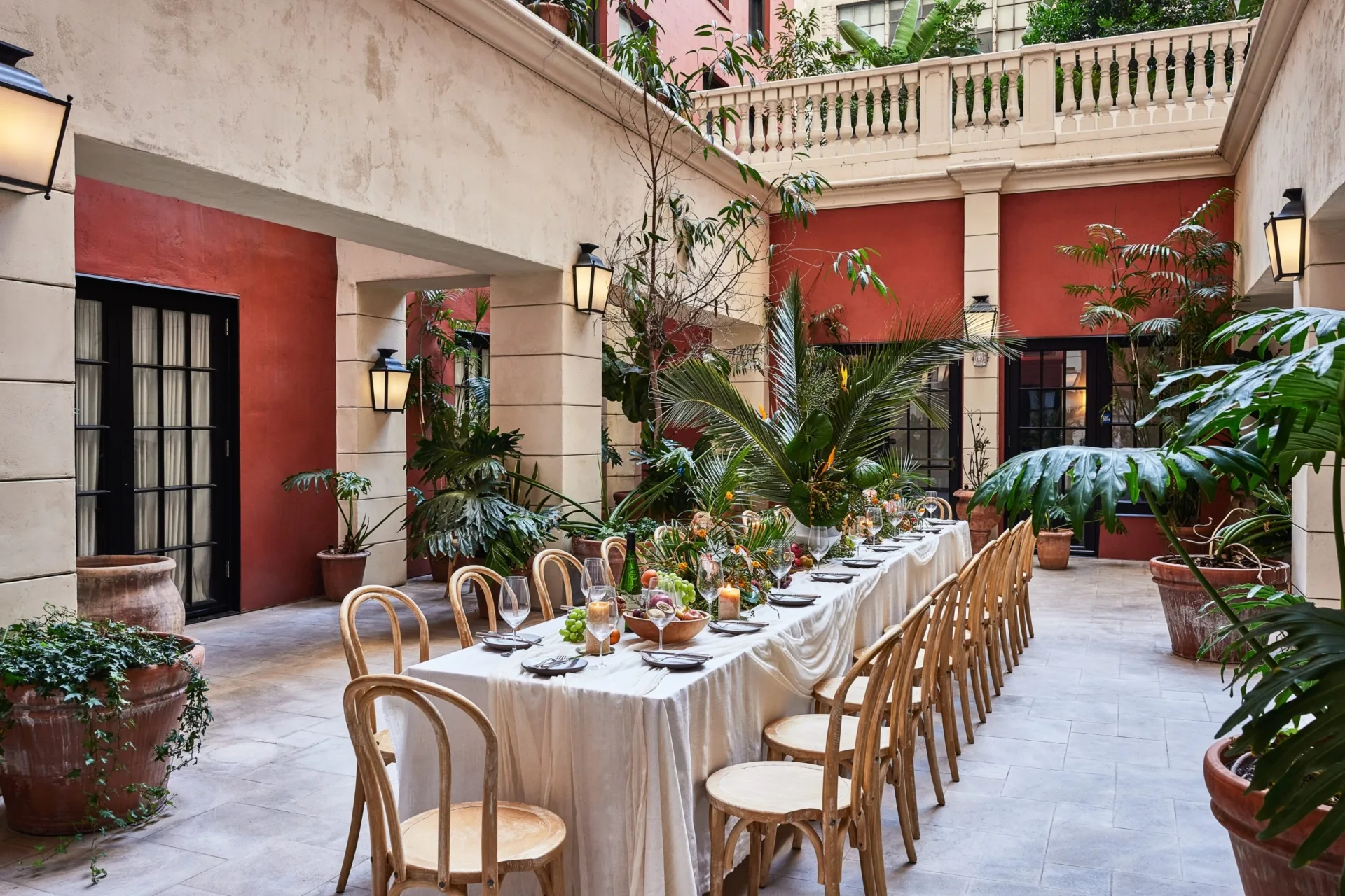 A long white banquet table is set for an outdoor dinner party in a lush, Mediterranean-style hotel courtyard.