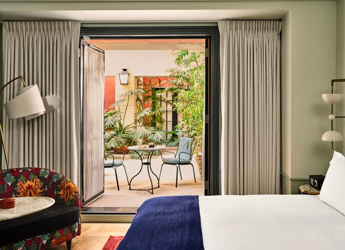 Sunlight fills a boutique hotel room at Hotel Per La, featuring a private patio with a table and leafy plants.
