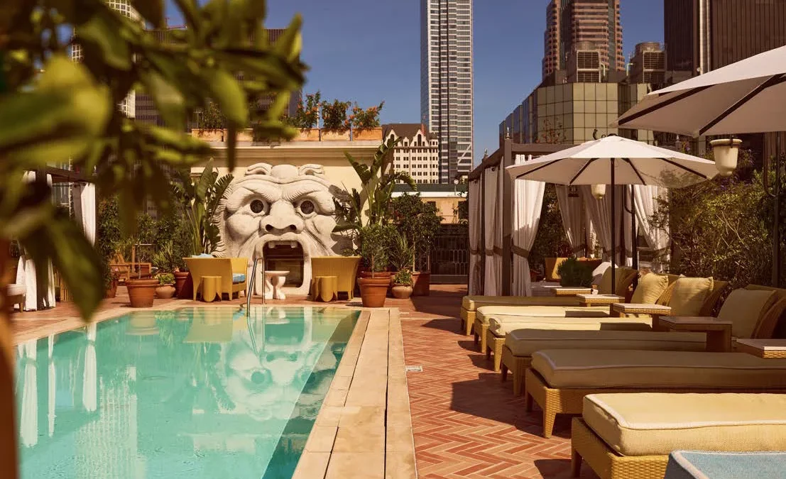 Sun loungers line a bright blue rooftop pool at Bar Clara with a sculpted stone face and city skyline views.