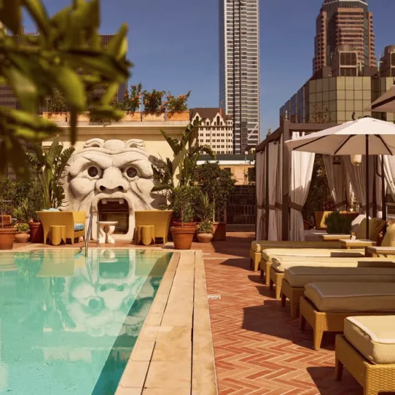 Sun loungers line a bright blue rooftop pool at Bar Clara with a sculpted stone face and city skyline views.