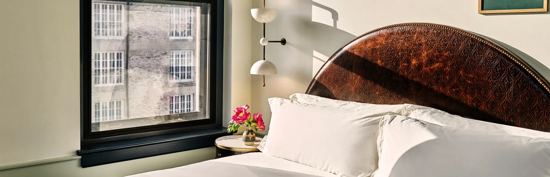 A sun-drenched hotel room at Hotel Per La features a detailed leather headboard and views of downtown Los Angeles.
