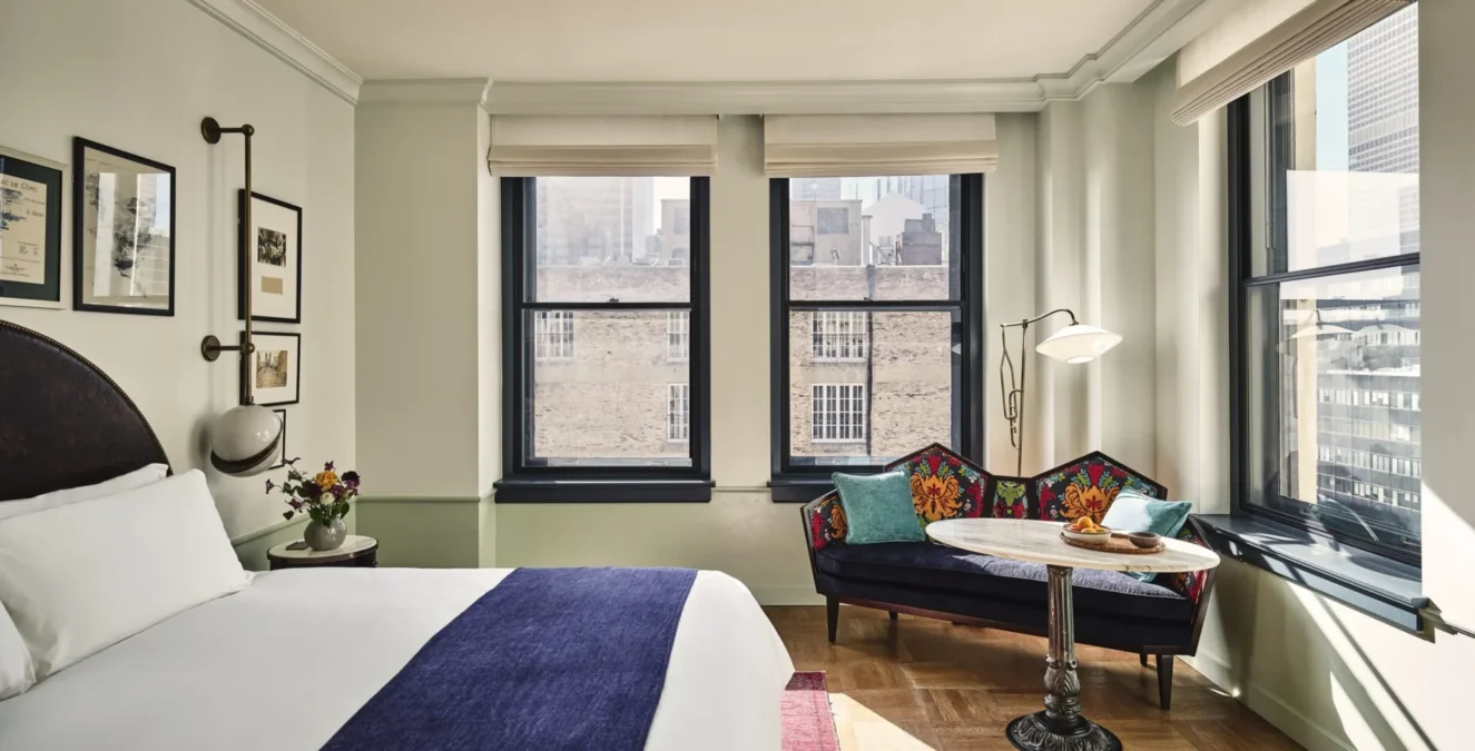 A stylish hotel room featuring a patterned settee, large windows, and plush bedding at Hotel Per La in Los Angeles.