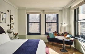 A stylish hotel room featuring a patterned settee, large windows, and plush bedding at Hotel Per La in Los Angeles.
