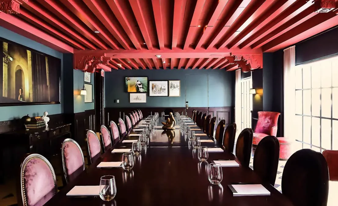 Pink velvet chairs surround a long wood table in a stylish boutique hotel meeting room with a red beamed ceiling.