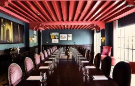 Pink velvet chairs surround a long wood table in a stylish boutique hotel meeting room with a red beamed ceiling.