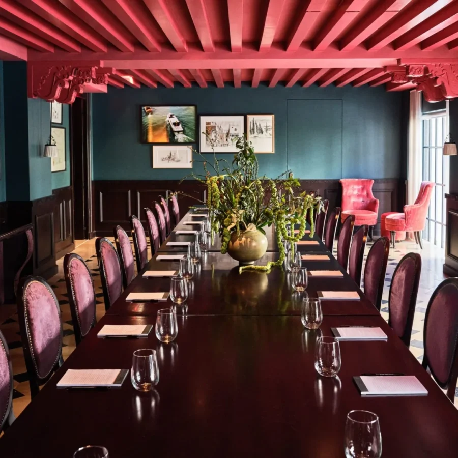 Velvet chairs surround a long dark wood table in a luxury Los Angeles event space with a red beamed ceiling.