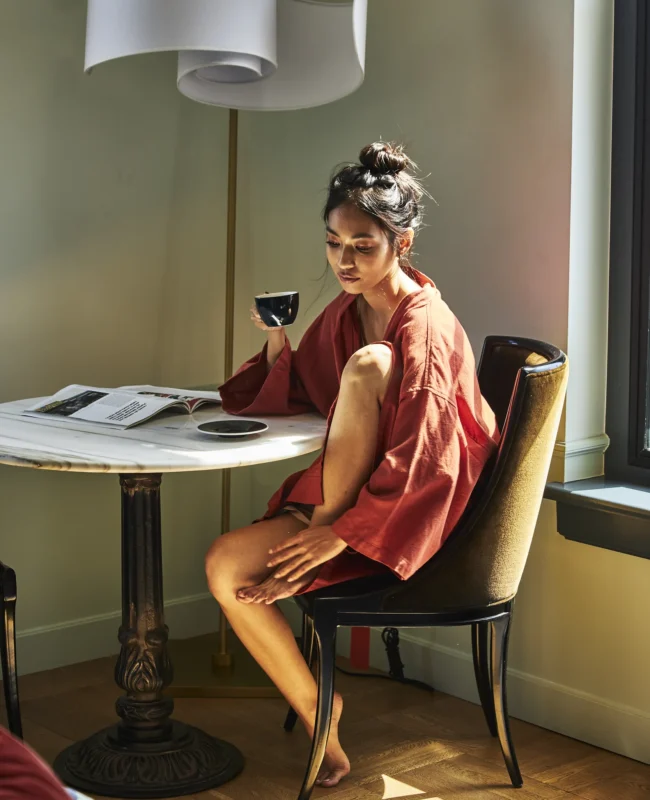 A woman in a red robe relaxes with coffee and a magazine in a sunlit boutique hotel room at Hotel Per La.