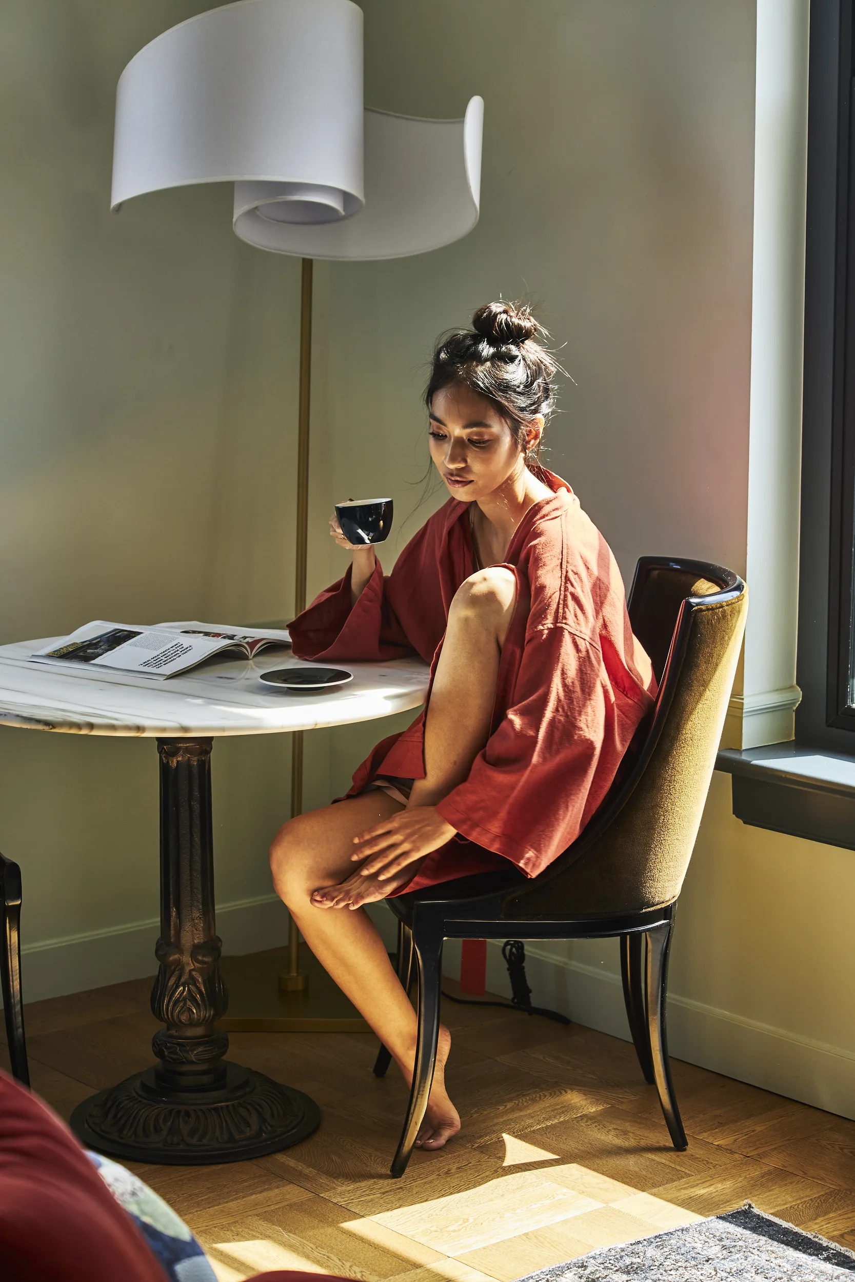 A woman in a red robe relaxes with coffee and a magazine in a sunlit boutique hotel room at Hotel Per La.