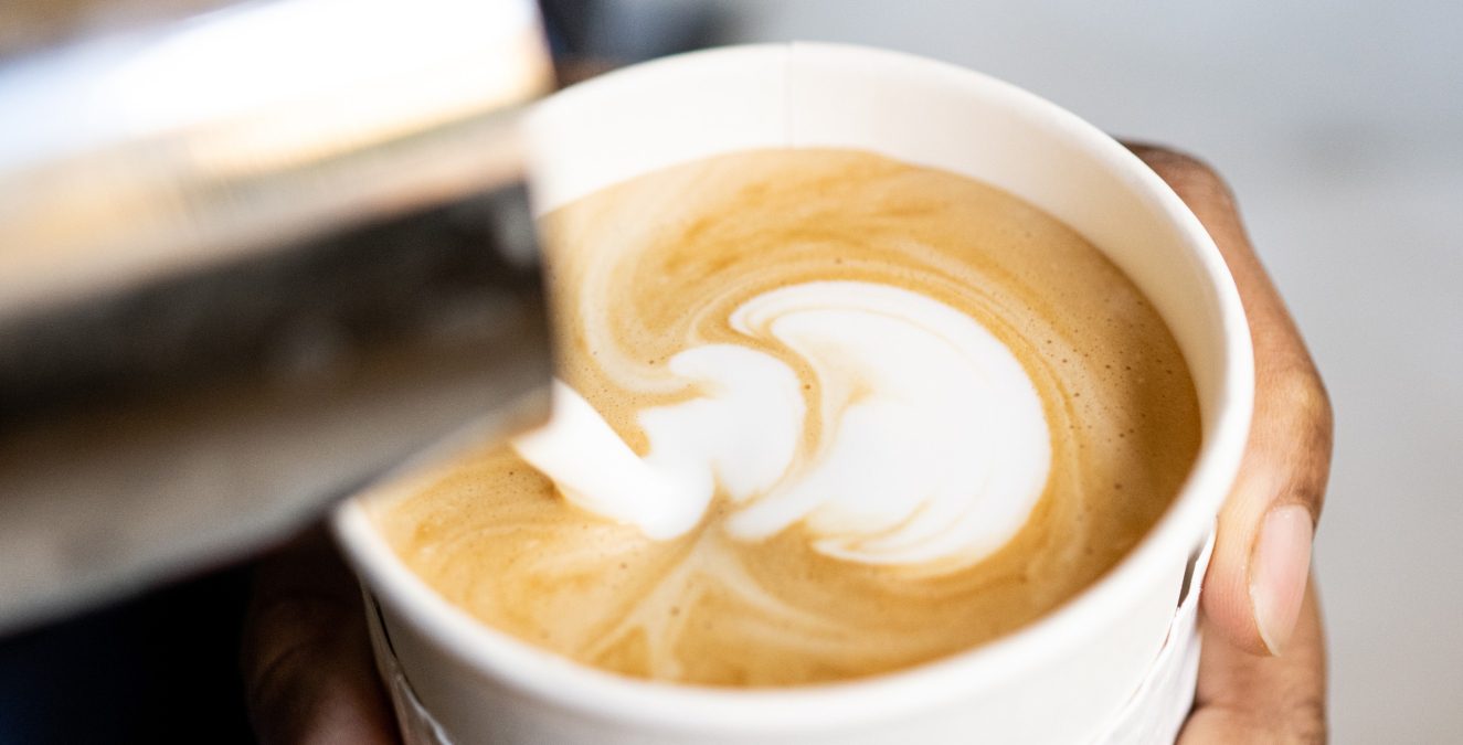 A barista pours steamed milk into a white paper cup with the Cafe Ora logo to create intricate latte art.