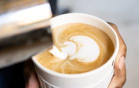 A barista pours steamed milk into a white paper cup with the Cafe Ora logo to create intricate latte art.