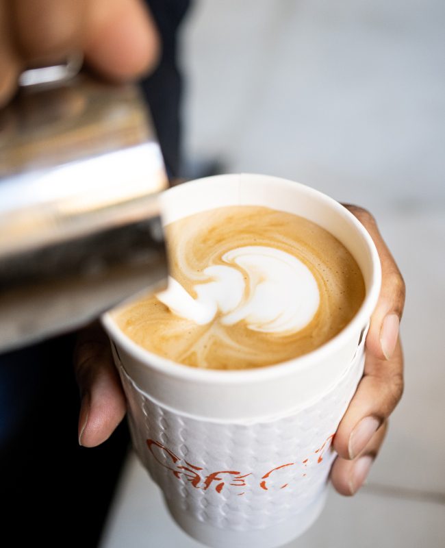 A barista pours steamed milk into a white paper cup with the Cafe Ora logo to create intricate latte art.