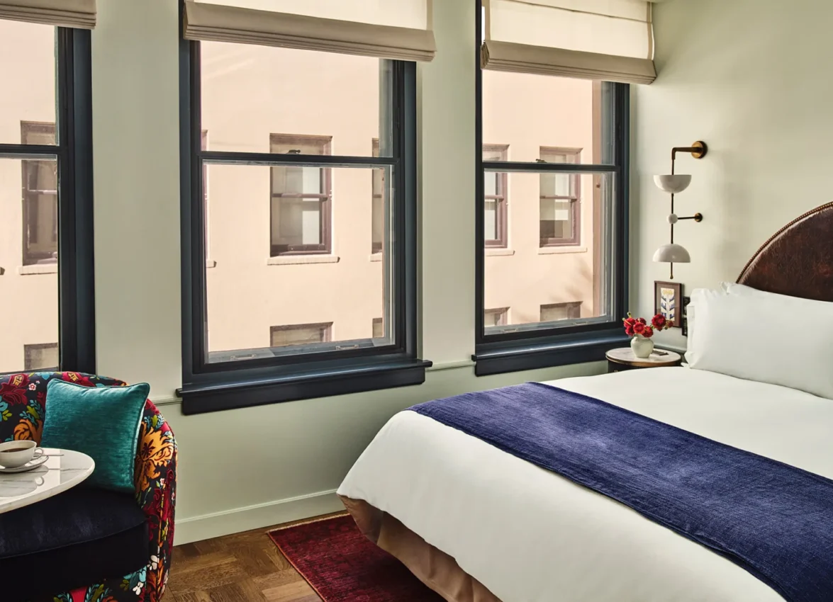 Sunlight streams into this Hotel Per La guest room featuring a plush bed and a colorful patterned armchair.