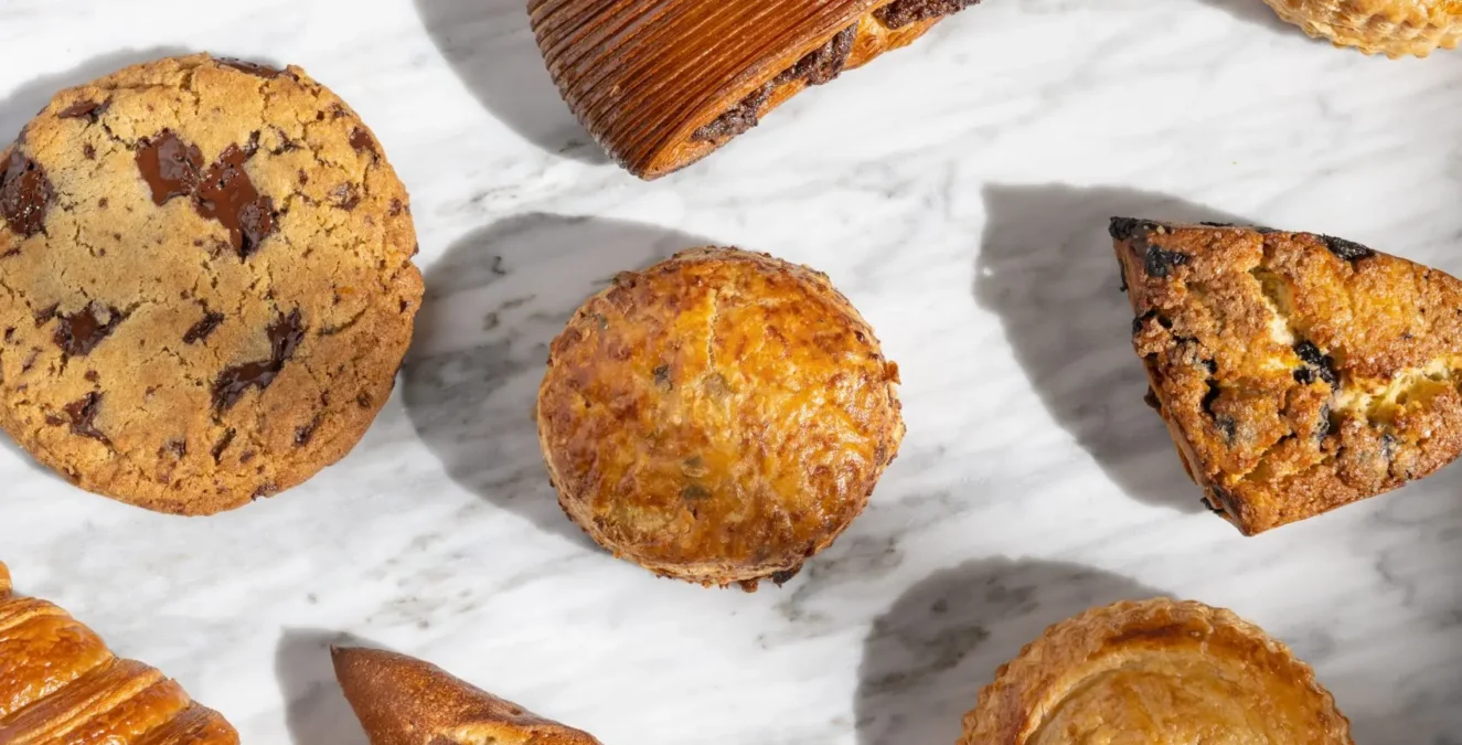 An assortment of golden-brown artisan pastries and fresh bread from Cafe Ora displayed on a white marble surface.