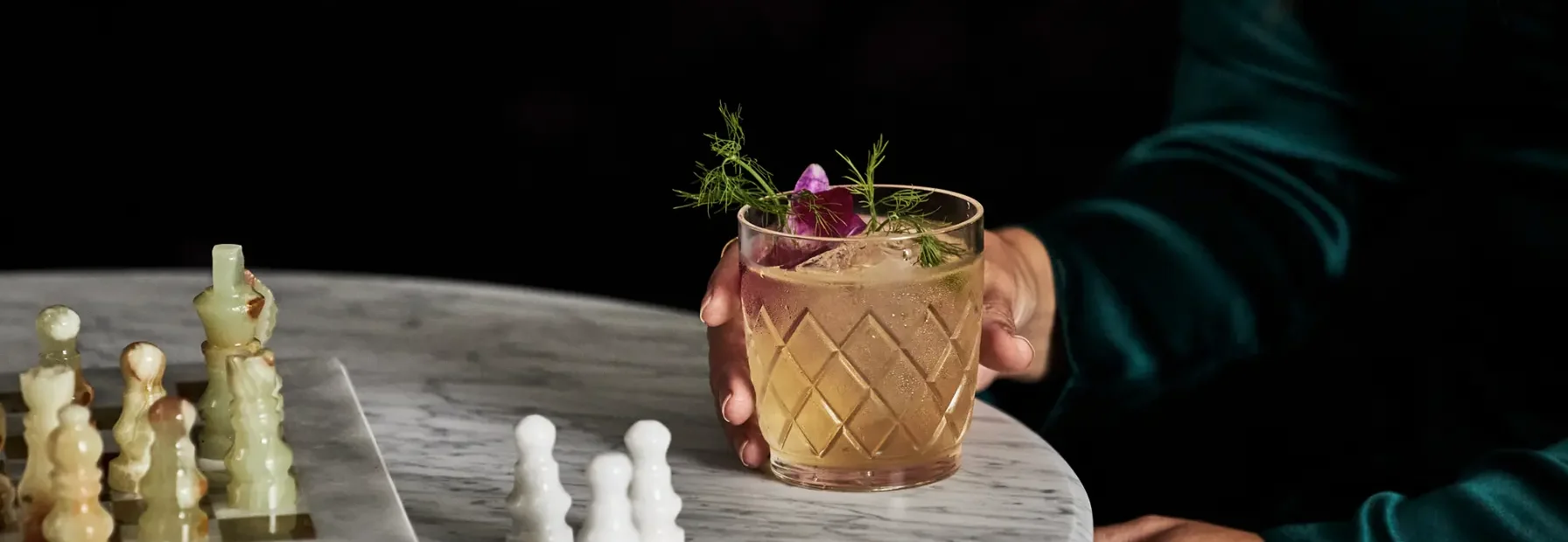 A person in a green dress holds a garnished cocktail beside a marble chess board at Hotel Per La in Los Angeles.
