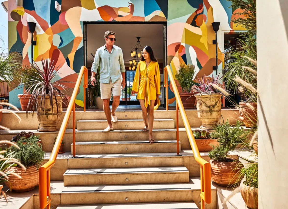 A couple walks down stone steps past vibrant murals and lush greenery at the Bar Clara rooftop bar in Los Angeles.