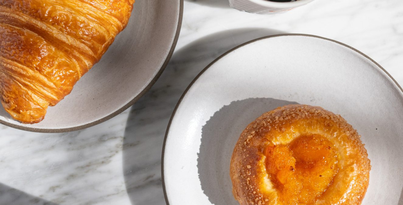Freshly baked croissant and fruit pastry served with a latte and black coffee on a marble table at Cafe Ora.