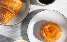 Freshly baked croissant and fruit pastry served with a latte and black coffee on a marble table at Cafe Ora.