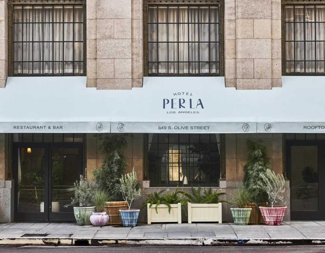 The street entrance of Hotel Per La features a white awning and potted plants along the downtown Los Angeles sidewalk.
