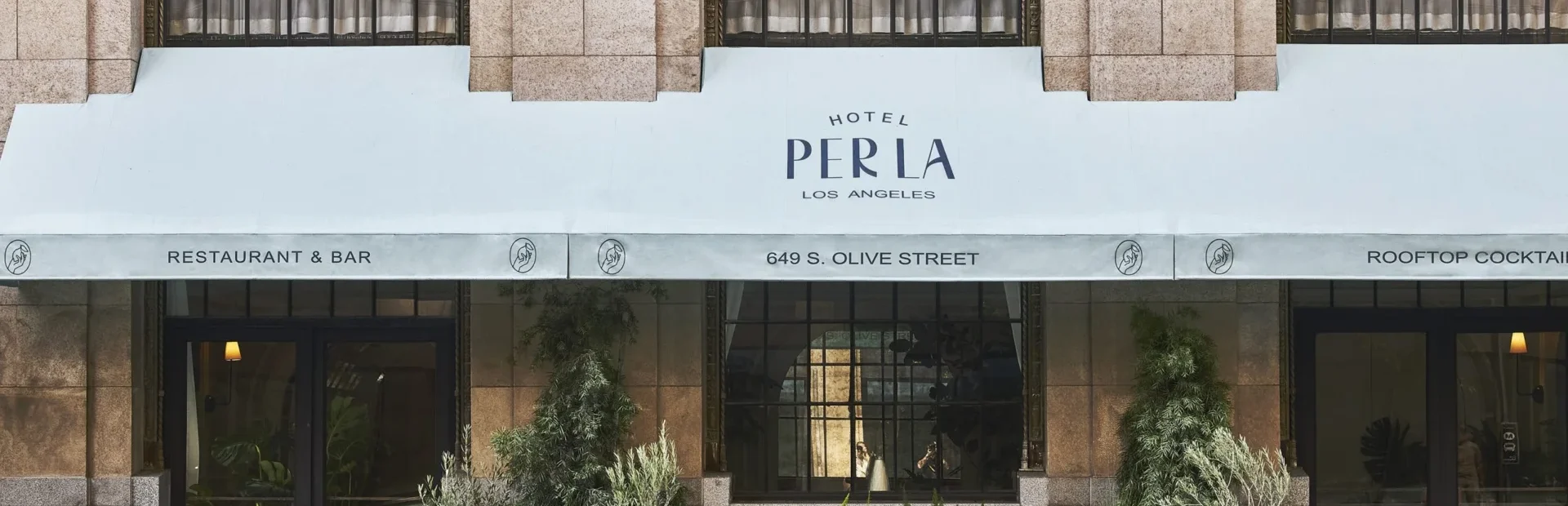 The street entrance of Hotel Per La features a white awning and potted plants along the downtown Los Angeles sidewalk.
