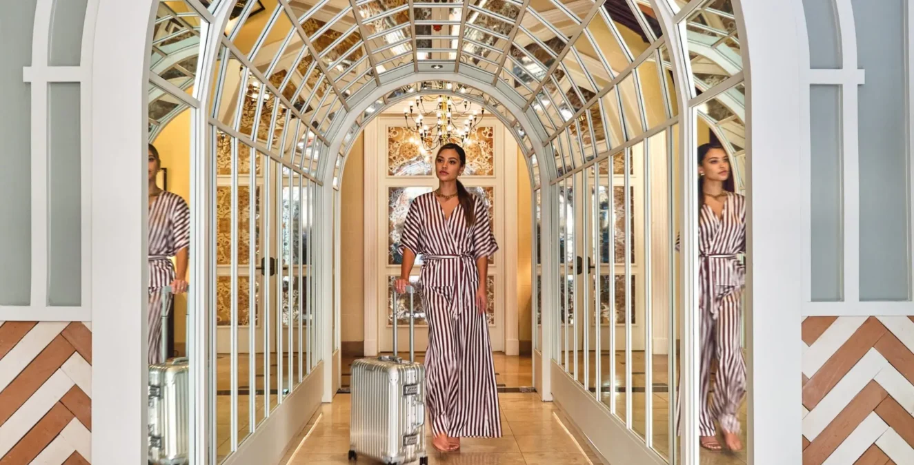 A guest pulls a silver suitcase through a grand mirrored arched hallway at a boutique hotel in Los Angeles.