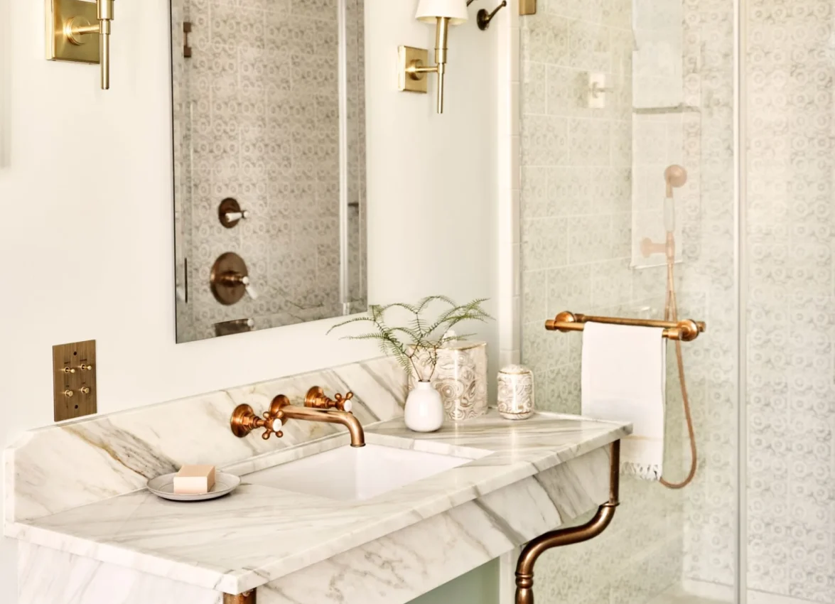 A marble vanity with gold fixtures and red flowers sits next to a glass-enclosed shower at Hotel Per La.