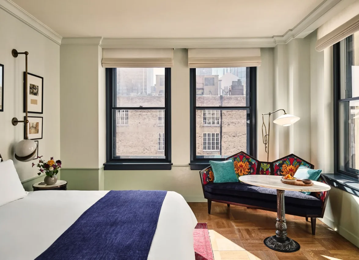 A stylish room at Hotel Per La with a blue bed runner, patterned sofa, and city views through large windows.