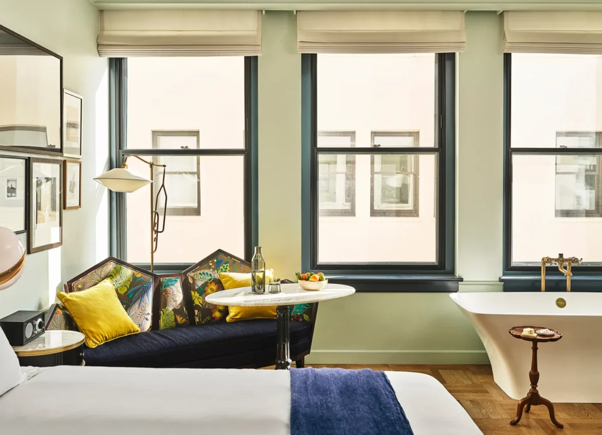 A sun-drenched hotel suite at Hotel Per La with a plush settee and a freestanding soaking tub by the window.