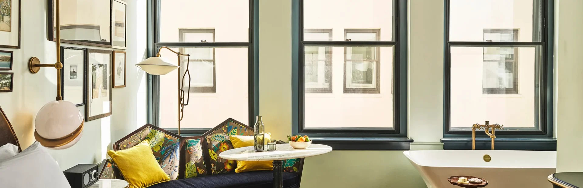 A sun-drenched hotel suite at Hotel Per La with a plush settee and a freestanding soaking tub by the window.