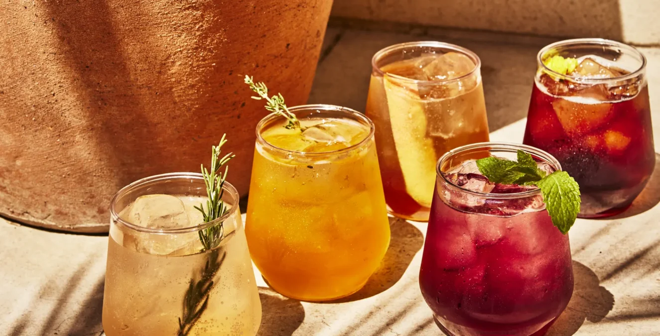 Five colorful cocktails with herb garnishes sit on a sunlit stone ledge at Bar Clara rooftop bar.