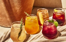 Five colorful cocktails with herb garnishes sit on a sunlit stone ledge at Bar Clara rooftop bar.