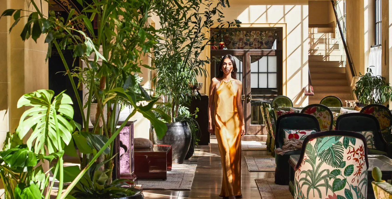 A woman in a gold silk dress walks through the sun-drenched, plant-filled lounge of Hotel Per La in Los Angeles.