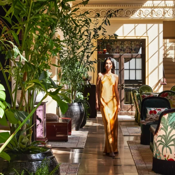 A woman in a gold silk dress walks through the sun-drenched, plant-filled lounge of Hotel Per La in Los Angeles.