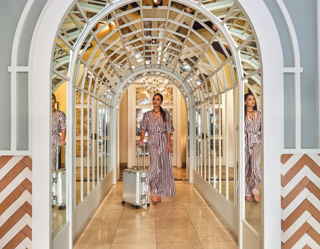 A guest pulls a silver suitcase through a striking mirrored arched hallway at Hotel Per La in Los Angeles.