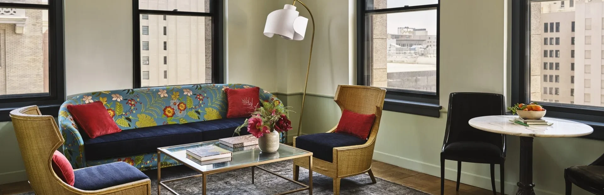 A vibrant living area at Hotel Per La with floral decor, wicker seating, and large windows overlooking downtown LA.