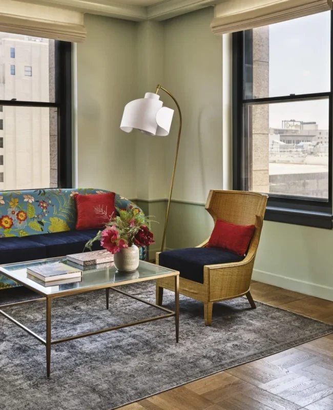 A vibrant living area at Hotel Per La with floral decor, wicker seating, and large windows overlooking downtown LA.