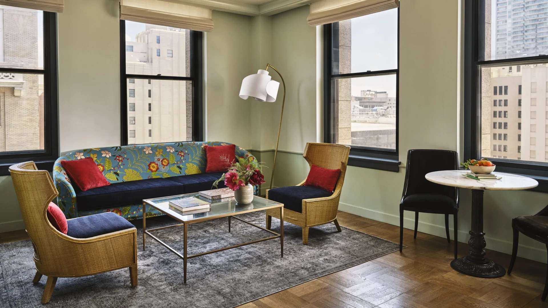 A vibrant living area at Hotel Per La with floral decor, wicker seating, and large windows overlooking downtown LA.