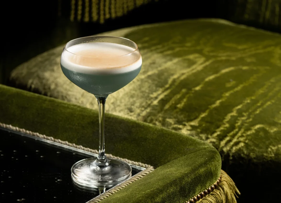 A chilled blue cocktail with foam rests on a table by plush green velvet at Bar Clara, a rooftop bar in Los Angeles.
