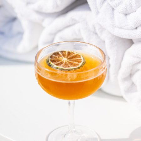 An amber cocktail with a dehydrated lime garnish rests on a white table next to rolled poolside towels.