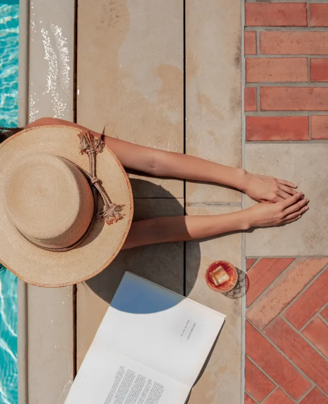 women in hat poolside with a book and drink,