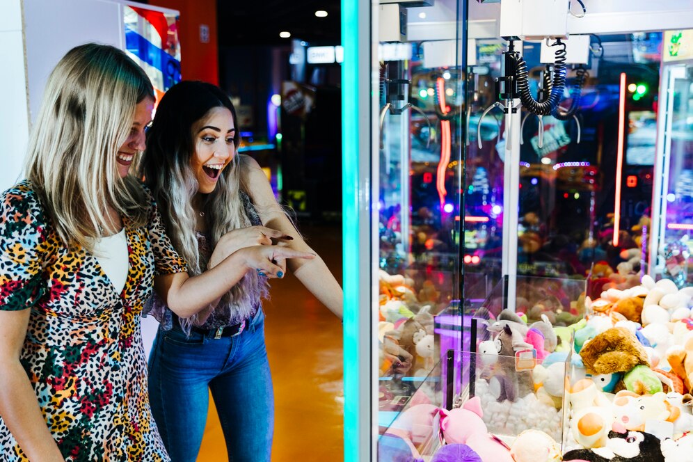 Two friends cheer as they try to win a prize from a claw machine at a bright arcade in downtown Los Angeles.