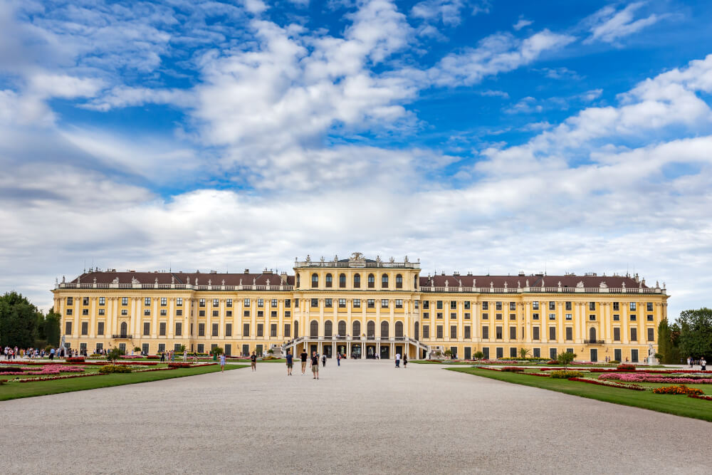 A majestic yellow palace with classical columns stands behind a wide gravel courtyard under a bright, cloudy sky.