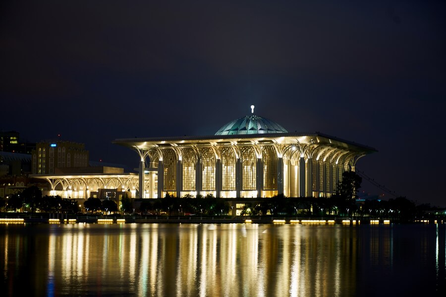 The glowing lights of a grand domed structure reflect across the calm surface of a lake at night.