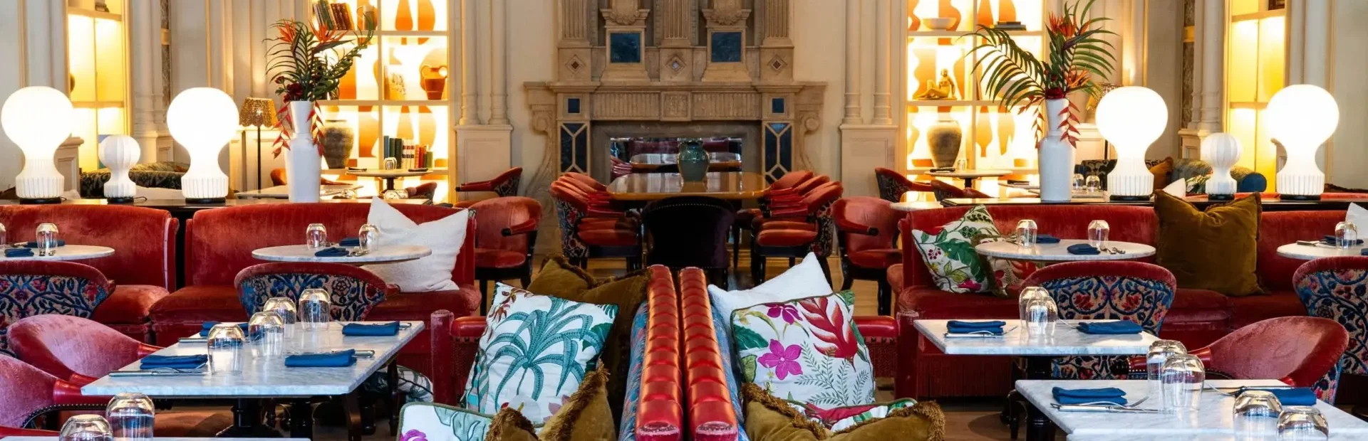 Luxurious restaurant interior featuring plush red velvet seating, vibrant floral and palm-patterned pillows, white marble tables set with glassware and blue napkins, and warm lighting from modern globe lamps. Ornate classical architecture and illuminated shelves with vases and decor accentuate the ambiance.