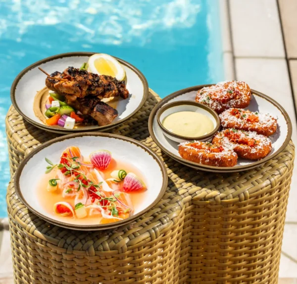 Three gourmet dishes served in white bowls on woven rattan pedestals beside a swimming pool. The dishes include grilled chicken skewers with lemon and vegetables, fresh sashimi or crudo garnished with radish slices and microgreens, and golden-brown fritters or cakes with a side of creamy dipping sauce. The setting shows beige pool deck tiles with turquoise pool water in the background
