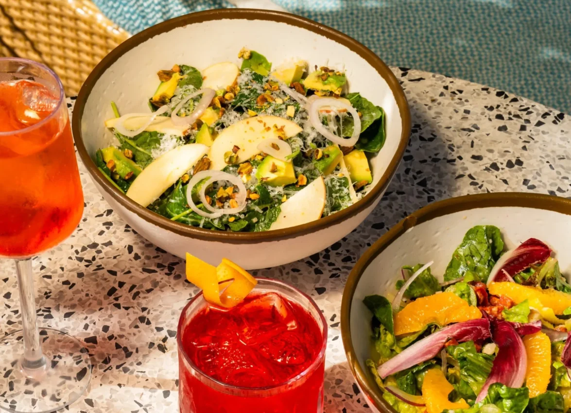 Two fresh salad bowls and red cocktails arranged on a speckled terrazzo table beside a swimming pool. The larger bowl contains a green salad with sliced onions, cheese, and mixed greens, while the smaller bowl features a citrus salad with orange segments and radicchio. Two red cocktails with orange garnishes complete the poolside dining setup, with turquoise pool water and golden woven furniture visible in the background.