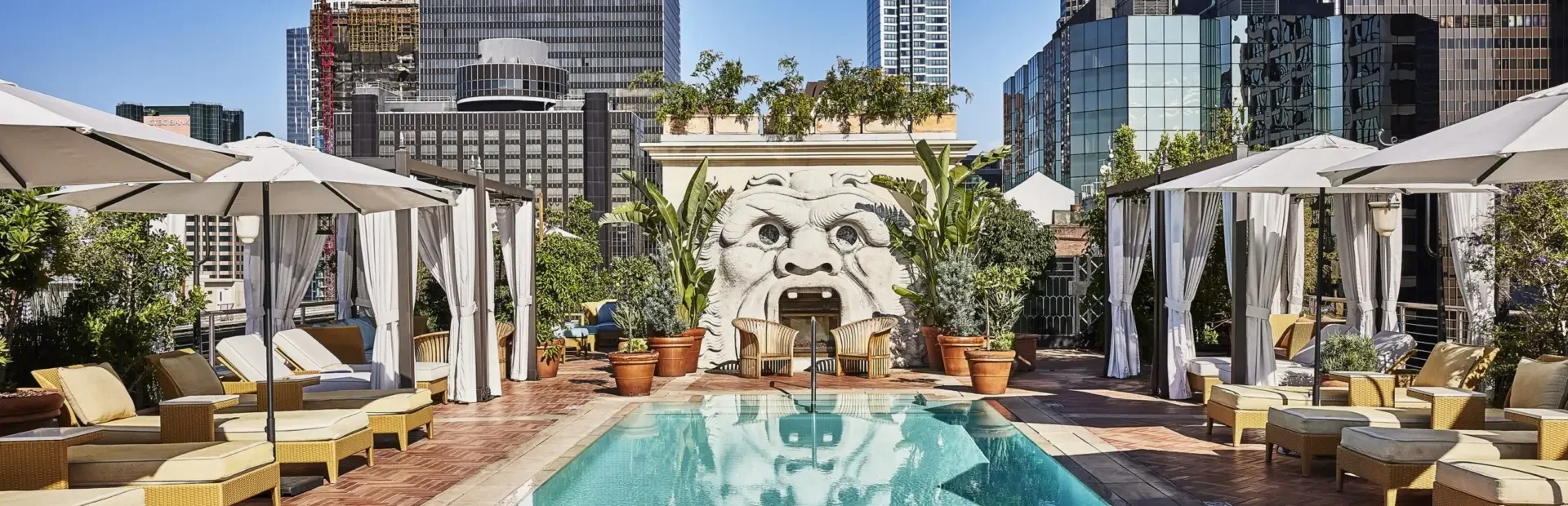 Luxurious rooftop pool deck with turquoise swimming pool surrounded by white cabanas and yellow wicker lounge chairs. A large sculptural face with an open mouth serves as a decorative water feature against the back wall, surrounded by potted plants. The downtown Los Angeles skyline with modern high-rise buildings rises in the background under a clear blue sky